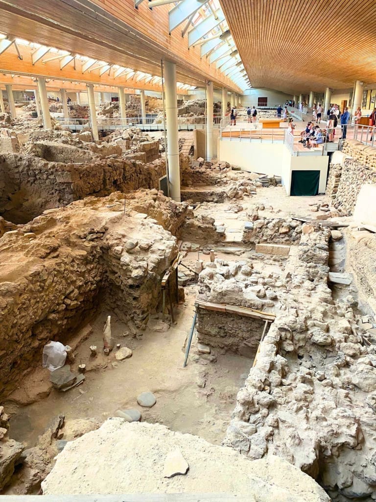 Santorini, Greece Travel Guide: Essential Tips for First-Time Visitors 9 Wide view of the Akrotiri Archaeological Site on Santorini, Greece, showing excavated ancient stone foundations and multi‑room structures preserved in a grid‑like pattern under a modern protective roof with wooden beams with walkways for visitors.