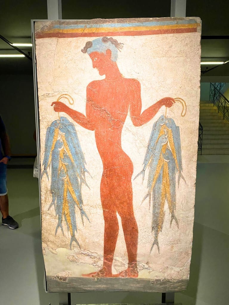 Santorini, Greece Travel Guide: Essential Tips for First-Time Visitors 13 “Fisherman Fresco” at the Museum of Prehistoric Thera in Santorini, Greece, depicting a stylized figure of a young man holding fish in both hands, painted in earthy red and blue tones on a light background.